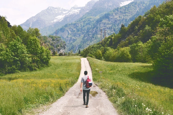 Fitness goals: A backpacker walks up a country road, flanked by greenery, as he makes his way toward mountains in the horizon.