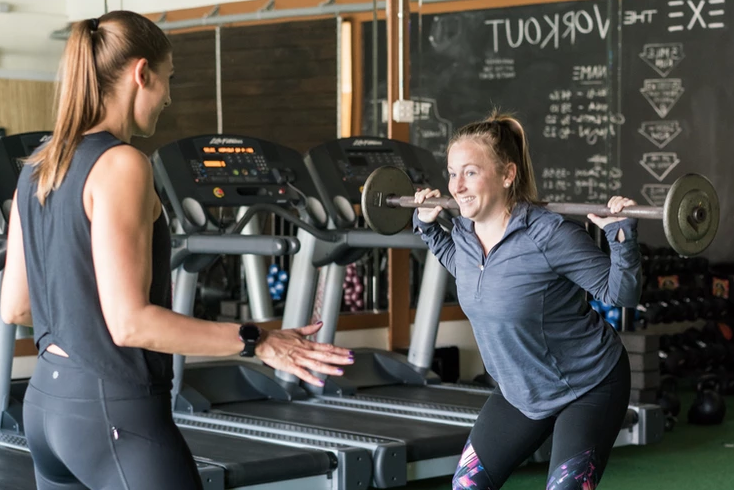 Breaking bad habits: This photo is of two women working out together. A brunette trainer faces a blond woman who is performing a squat exercise.