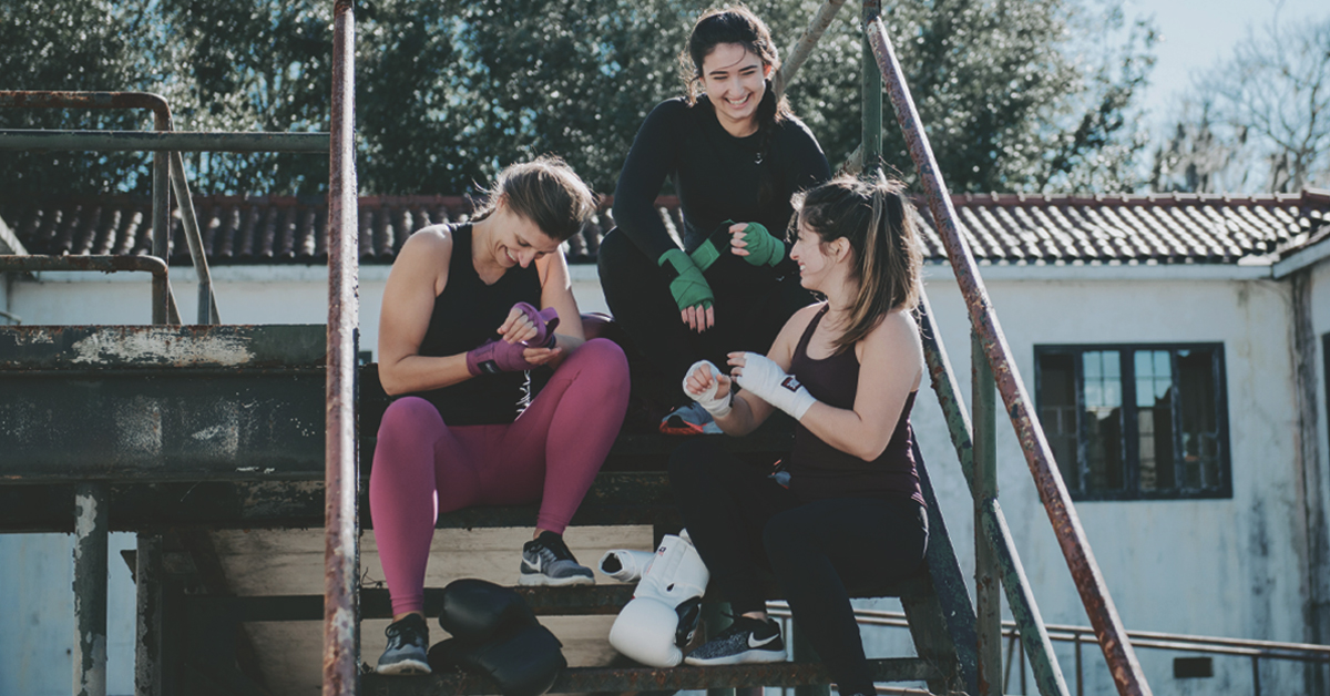Training with FYS: Three women wearing workout attire sit on a short flight of steps outside and smile during an engaging conversation.